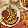 A bottle of Cholula Original Mexican Hot Sauce 150Ml sits next to a plate of crispy birria tacos served with a side of consomme and fresh lime wedges.