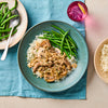 A rustic blue bowl holds a serving of creamy beef stroganoff with mushrooms and onions over white rice, next to a pile of steamed green beans.