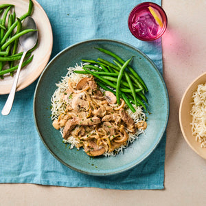 A rustic blue bowl holds a serving of creamy beef stroganoff with mushrooms and onions over white rice, next to a pile of steamed green beans.