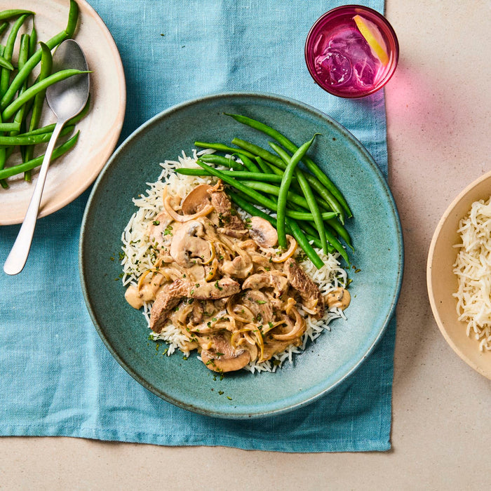 A rustic blue bowl holds a serving of creamy beef stroganoff with mushrooms and onions over white rice, next to a pile of steamed green beans.