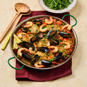 A large paella pan is filled with a colorful rice dish containing chicken, shrimp, mussels, and peas, served with lemon wedges and a side salad.