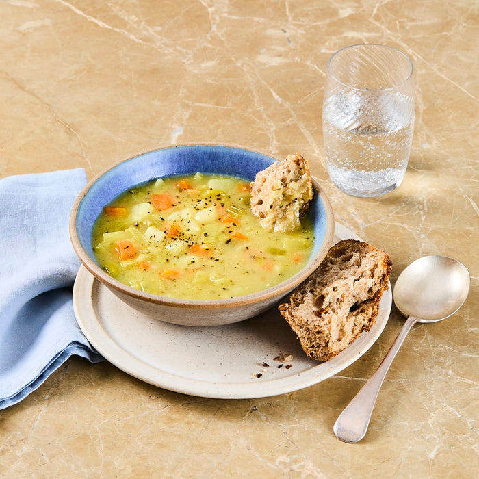 A comforting bowl of chunky vegetable soup, seasoned with black pepper, is served on a marble countertop with a piece of crusty bread for dipping and a glass of sparkling water.