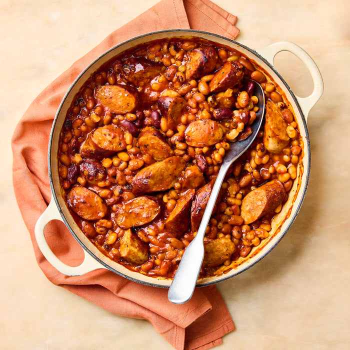 An overhead shot of a hearty sausage and bean casserole in a white enameled pot with a serving spoon.