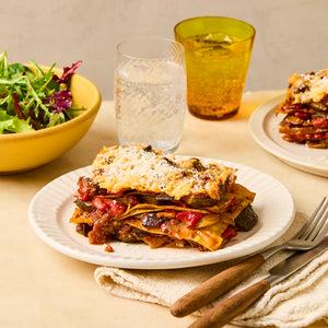 A slice of vegetable lasagna sits on a white plate, served with a side salad and a glass of sparkling water.