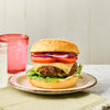 A juicy cheeseburger with lettuce, tomato, and onion on a sesame seed bun is served on a plate with drinks in the background.
