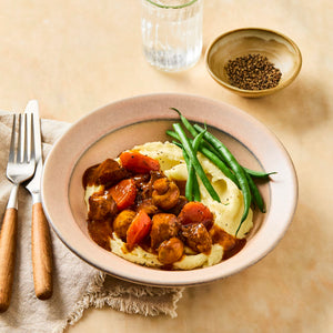 A comforting meal of beef stew with carrots and mushrooms is served over creamy mashed potatoes with a side of green beans, with a small bowl of cracked black pepper nearby.