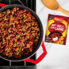 A red pot filled with freshly made chilli sits next to a wooden spoon and a packet of Schwartz Hot Chilli Con Carne Recipe Mix 41G on a marble countertop.