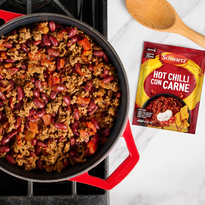A red pot filled with freshly made chilli sits next to a wooden spoon and a packet of Schwartz Hot Chilli Con Carne Recipe Mix 41G on a marble countertop.