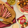 A serving suggestion for Schwartz Creamy Pepper Sauce Mix 25G shows sliced steak and roasted potatoes on a plate and cutting board, with a bowl of the finished sauce.