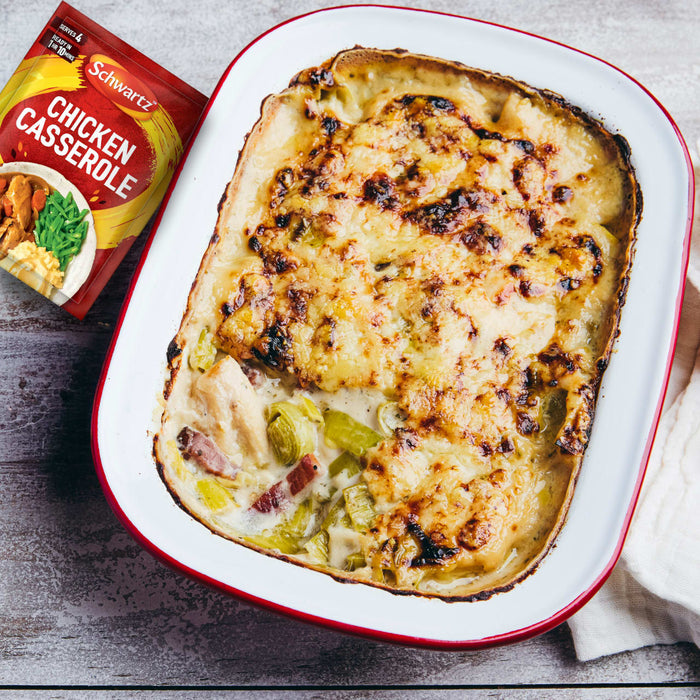 A freshly baked chicken and leek casserole with a golden-brown cheesy topping is presented in a white and red baking dish, next to the packet of Schwartz Chicken Casserole Recipe Mix 36G used to prepare the meal.