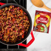 A red pot filled with freshly made chilli con carne sits on a stovetop, with a packet of Schwartz Chilli Con Carne Recipe Mix 41G and a wooden spoon resting on the counter beside it.
