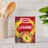 A sachet of Schwartz Lasagne Recipe Mix 36G is placed on a kitchen countertop, with cooking utensils and a plant in the background to create a home-cooking scene.