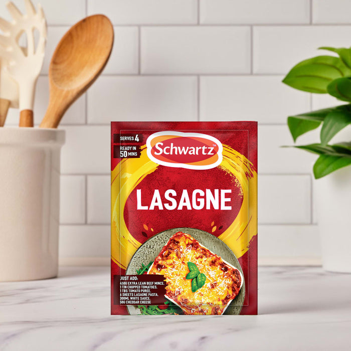 A sachet of Schwartz Lasagne Recipe Mix 36G is placed on a kitchen countertop, with cooking utensils and a plant in the background to create a home-cooking scene.