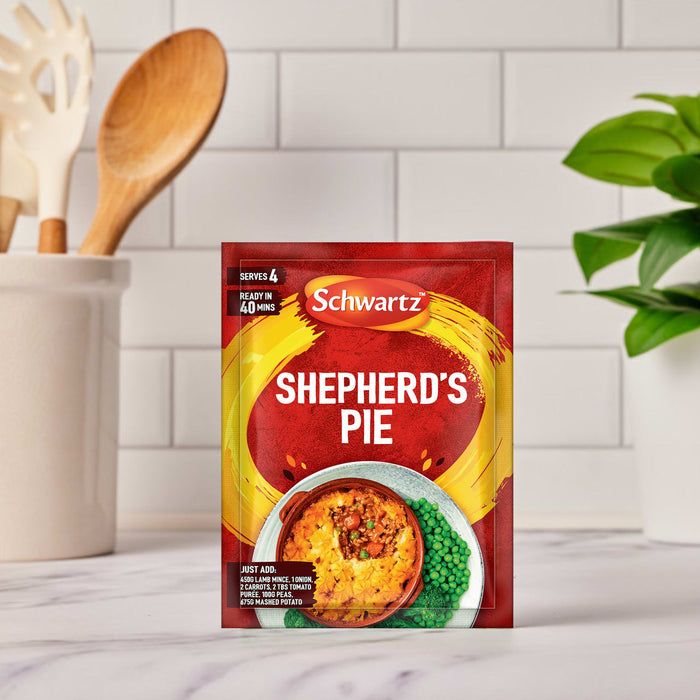 A packet of Schwartz Shepherd'S Pie Recipe Mix 38G is set on a kitchen counter, with cooking utensils and a plant blurred in the background.