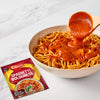 A wooden spoon pours a rich bolognese sauce over a large bowl of spaghetti, with a packet of Schwartz Spaghetti Bolognese Recipe Mix 40G displayed in the foreground to show the finished meal.