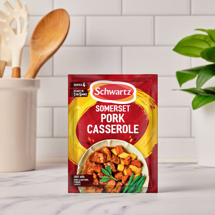 A packet of Schwartz Somerset Pork Casserole Recipe Mix 36G is displayed on a kitchen countertop, with a utensil holder and a green plant blurred in the background.