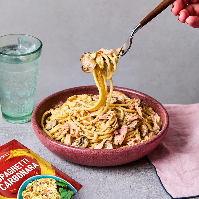 A delicious bowl of spaghetti carbonara, made using a packet of Schwartz Spaghetti Carbonara Recipe Mix 32G, is being twirled on a fork next to a glass of sparkling water.