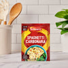 A packet of Schwartz Spaghetti Carbonara Recipe Mix 32G is displayed on a marble kitchen counter, with a utensil holder and a green plant out of focus in the background.