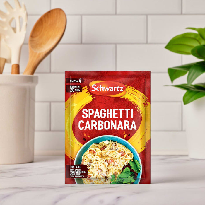 A packet of Schwartz Spaghetti Carbonara Recipe Mix 32G is displayed on a marble kitchen counter, with a utensil holder and a green plant out of focus in the background.