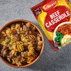 A brown bowl filled with a creamy beef and fusilli pasta dish is presented alongside a packet of Schwartz Beef Casserole Recipe Mix 38G against a grey, textured background.