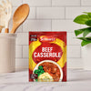 A packet of Schwartz Beef Casserole Recipe Mix 38G sits on a kitchen counter, with a utensil holder and a small plant visible in the blurred background.