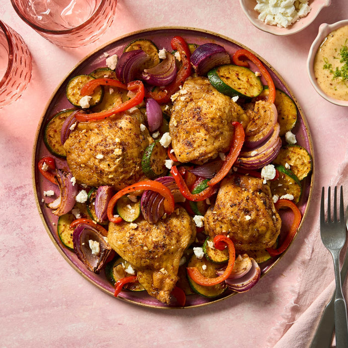 Roasted chicken thighs and colorful vegetables are seasoned to perfection with Schwartz Cajun Seasoning 110G and served on a pink plate with a side of feta cheese and creamy dip.