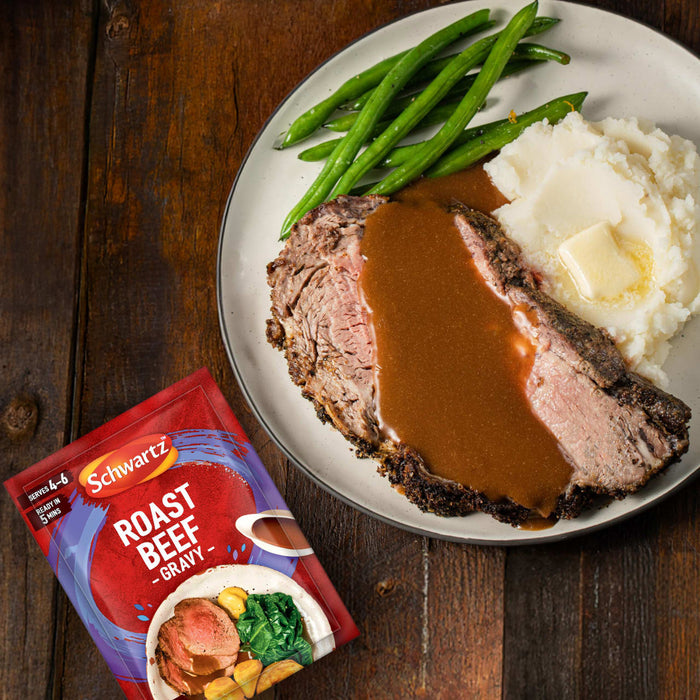 A finished meal of roast beef with gravy, mashed potatoes, and green beans is displayed on a plate, with a packet of Schwartz Roast Beef Gravy Mix 27G placed next to it on a wooden surface.