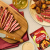 A delicious meal of sliced steak and roasted potatoes is served alongside a packet of Schwartz Creamy Mild Peppercorn Sauce Mix 25G, which can be used to create the accompanying creamy sauce.