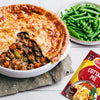 A delicious meat pie with a golden puff pastry crust, served with green beans and peas, is displayed next to the Schwartz Sa Cottage Pie seasoning packet used to make the filling.