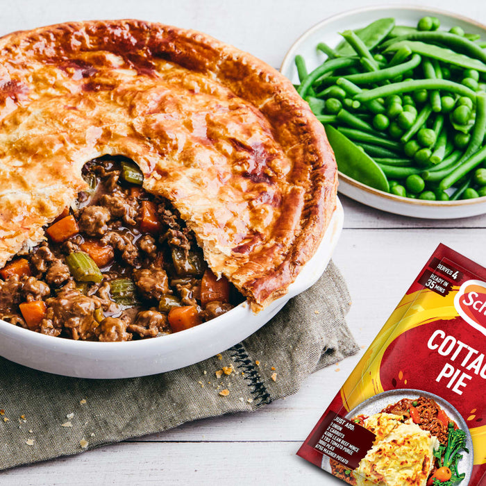 A delicious meat pie with a golden puff pastry crust, served with green beans and peas, is displayed next to the Schwartz Sa Cottage Pie seasoning packet used to make the filling.