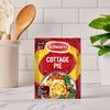 A packet of Schwartz Sa Cottage Pie recipe mix is displayed on a kitchen counter, with a utensil holder and a plant blurred in the background.