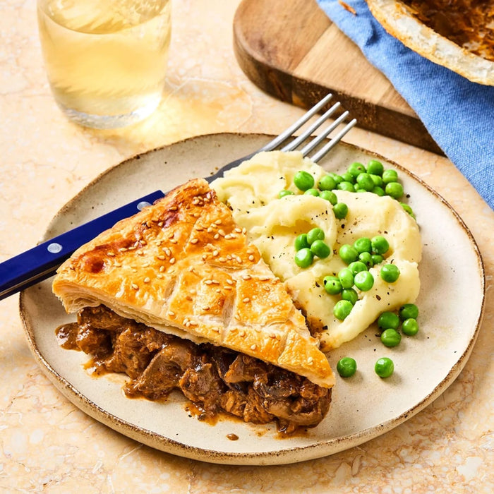 A golden-brown slice of Sa Cottage Pie with a flaky puff pastry crust is served on a rustic plate with creamy mashed potatoes and green peas.
