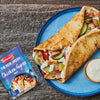 A delicious-looking chicken gyros, filled with chicken, vegetables, and tzatziki sauce, is served on a blue plate next to a packet of Schwartz The Real Greek Chicken Gyros Wrap 20G seasoning mix, which was used to prepare the meal.