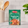 A packet of Schwartz Bella Italia Gamberoni Garlic & Chilli Prawn Spaghetti 35G sits on a kitchen countertop, with cooking utensils and a plant blurred in the background.