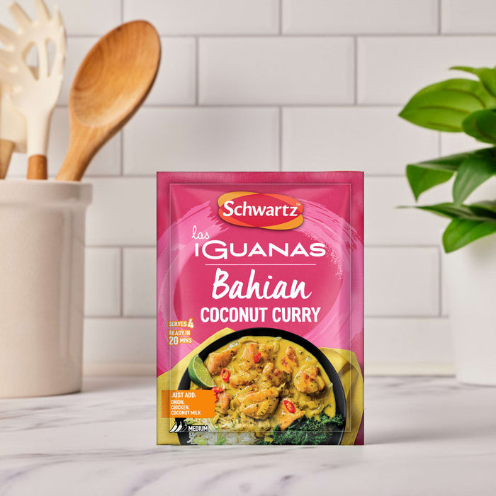 A vibrant pink packet of Schwartz Las Iguanas Bahian Coconut Curry 25G is displayed on a kitchen counter, with a serving suggestion of the finished dish shown on the front.