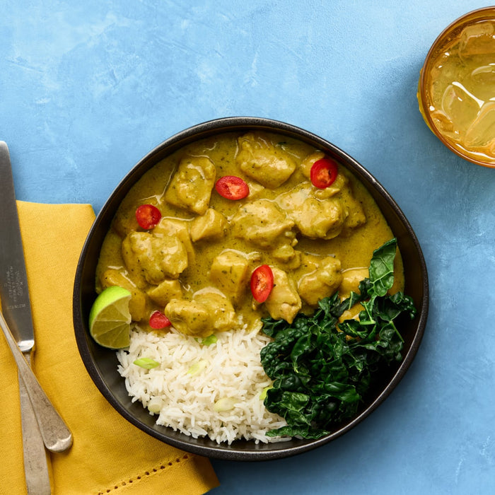 A delicious bowl of Bahian coconut curry, made with Schwartz Las Iguanas Bahian Coconut Curry 25G, is served with rice, kale, and a lime wedge on a blue surface.