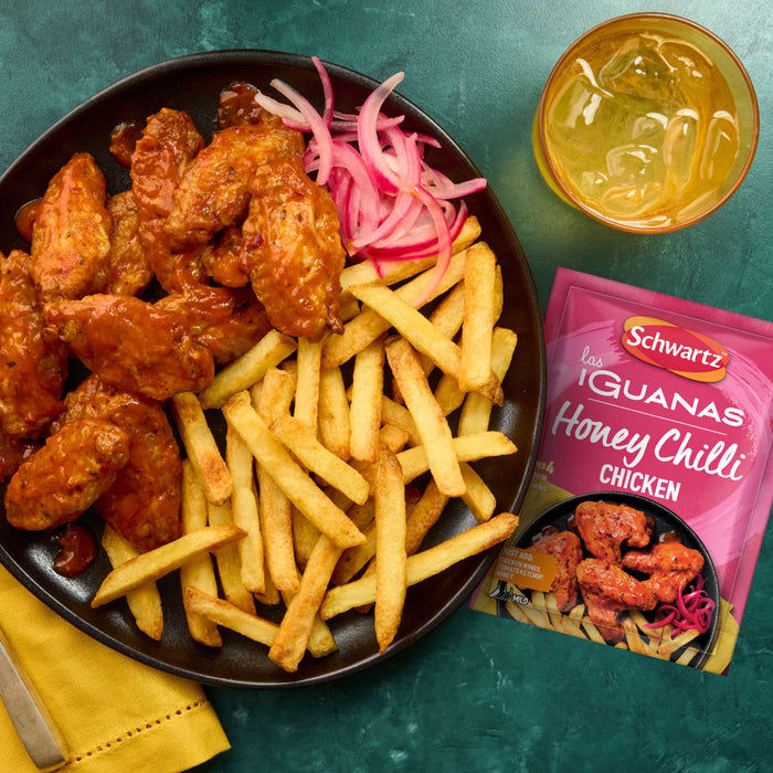 A plate of crispy fries and chicken wings coated in a sweet and spicy glaze is presented alongside a packet of Schwartz Las Iguanas Honey Chilli Chicken 35G and a refreshing iced drink.