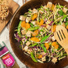 A vibrant Asian-style salad featuring tofu, mandarin oranges, and snap peas is presented in a dark wooden bowl alongside a bottle of Schwartz Chinese 5 Spice Seasoning 58G.