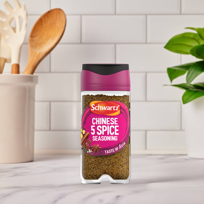 A jar of Schwartz Chinese 5 Spice Seasoning 58G stands on a marble countertop in a bright kitchen setting with wooden utensils and a green plant in the background.