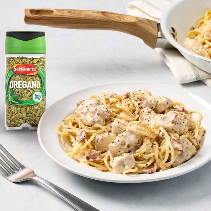 A jar of Schwartz Oregano 7G is placed next to a white plate of creamy chicken and bacon spaghetti, which has been sprinkled with the herb, with a pan of the pasta in the background.