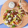 Two delicious gyros, whose meat is seasoned with Schwartz Oregano 7G, are served on a wooden platter with a side of tzatziki sauce.