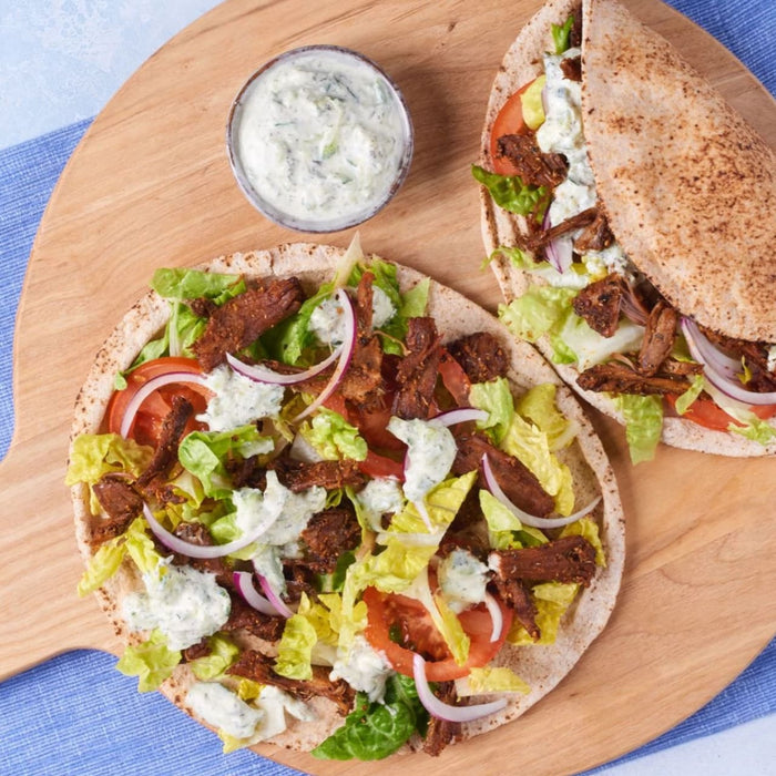 Two delicious gyros, whose meat is seasoned with Schwartz Oregano 7G, are served on a wooden platter with a side of tzatziki sauce.
