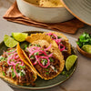 A plate of delicious shredded meat tacos, seasoned with spices like Schwartz Oregano 7G, are topped with pickled red onions, cilantro, and crema, and served with lime wedges.