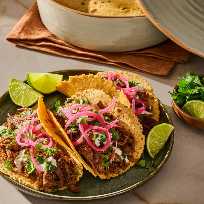 A plate of delicious shredded meat tacos, seasoned with spices like Schwartz Oregano 7G, are topped with pickled red onions, cilantro, and crema, and served with lime wedges.
