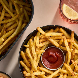 A top-down view of golden crispy french fries seasoned with Schwartz Af Fries Seas served in a black bowl with a side of ketchup and a refreshing lemon water.