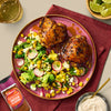 Two roasted chicken thighs seasoned with Schwartz Paprika and Onion Chicken Seasoning 50G are served on a pink plate alongside a vibrant corn and avocado salad.