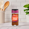 A bottle of Schwartz Paprika & Onion Chicken Seasoning 50G stands on a marble countertop in a bright kitchen setting with wooden utensils and a green plant in the background.