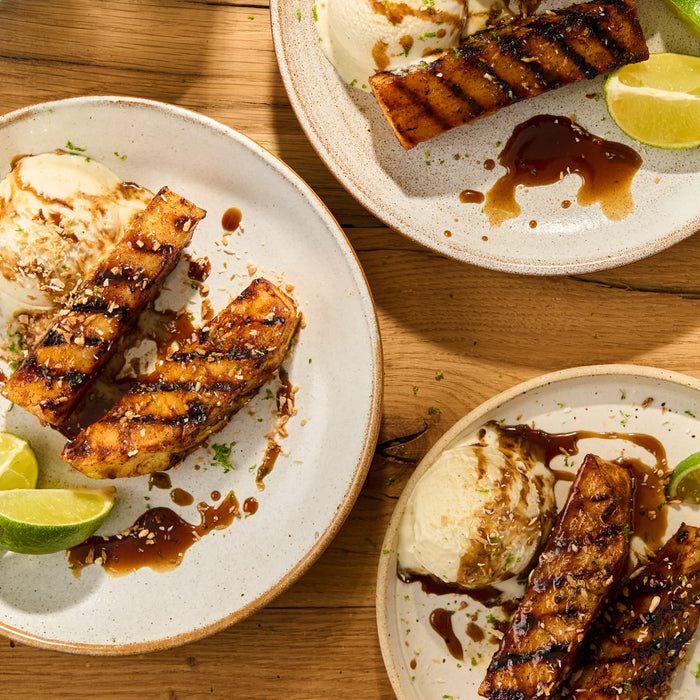 Grilled pineapple spears, seasoned with Schwartz Ground Cinnamon 39G, are served on rustic plates with vanilla ice cream, a dark sauce, and fresh lime.