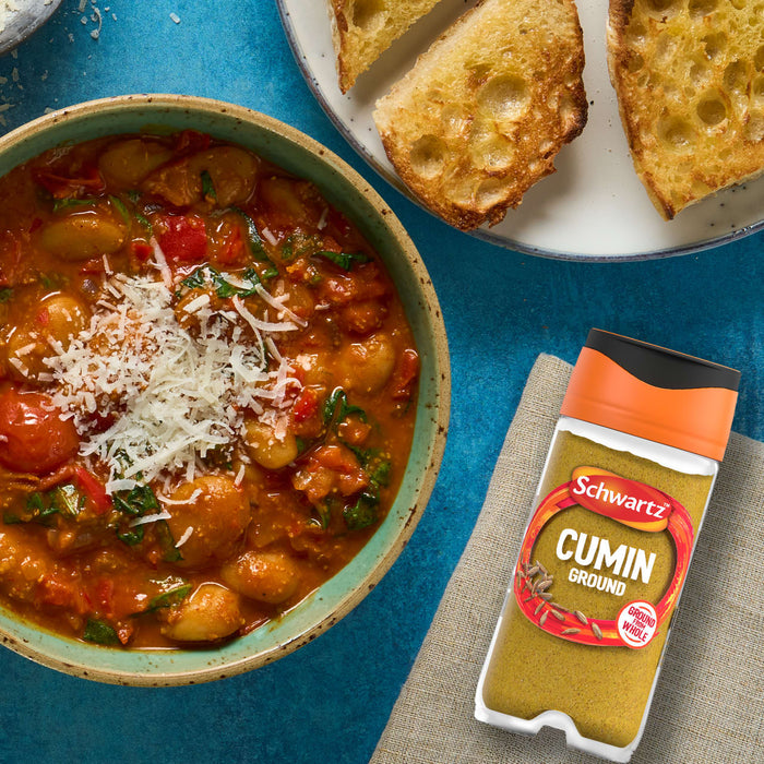 A jar of Schwartz Cumin Ground 37G sits next to a rustic bowl of bean and tomato stew, which is topped with grated cheese and served with toasted bread.
