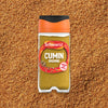 A clear jar of Schwartz Cumin Ground 37G is centered on a textured background of the ground spice.